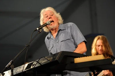 John Mayall singing into a microphone and playing a keyboard on stage.