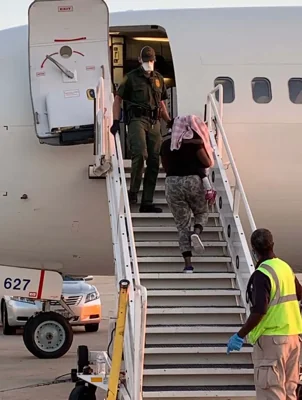 People walk up a mobile staircase to board a repatriation flight on a runway while US immigration officials look on from the top and bottom of the stairs.