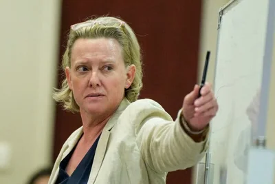 A woman with glasses in her hair holds a pen with her left hand near a whiteboard.