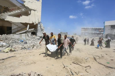 people carry a body wrapped in white plastic past destroyed buildings and sand/dust rising