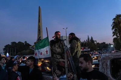 A Syrian rebel fighter, dressed in camouflage, stands next to an opposition flag, surrounded by cars and other people celebrating the overthrow of the Assad dictatorship.