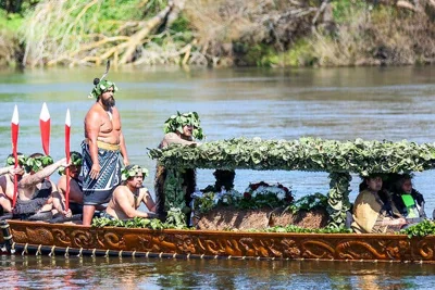 People sit and stand in an elaborately carved wooden. They are wearing crowns made of leaves, and some sit beneath a canopy also made of leaves.