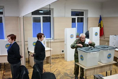 An older man wearing camouflage can be seen in a mirror placing his ballot into a plastic box. A woman stands monitoring him, and is also reflected in the mirror.