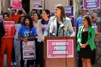 Stephanie Stahl Hamilton, an Arizona lawmaker, speaking at a lectern with a sign on it reading “Democrats Protect Abortion.” Abortion-rights activists stand behind her holding signs.