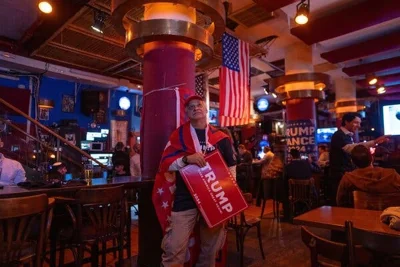 A person holds a Trump flier in a bar decorated with American flags and a Trump-Vance poster.