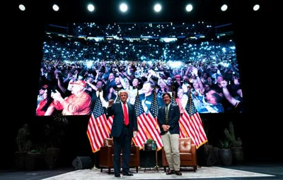 Donald Trump, left, and Tucker Carlson, right, stand on a stage with at least five American flags behind them. Behind the flags is a screen showing the crowd at the event.