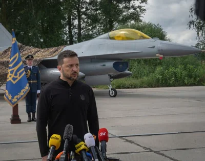 President Volodymyr Zelensky speaking in front of microphones outside with a military jet in the background.