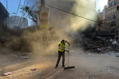 A worker cleans a street as smoke rises from a destroyed building that was hit by an Israeli airstrike in Dahiyeh earlier on Sunday