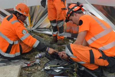 Three rail workers, one squatting, one sitting and one standing, partially in frame, wearing orange high-visibility gear.