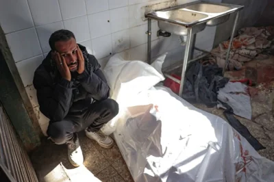 A Palestinian man sits next to the body of a person killed in an Israeli strike inside Al-Aqsa Martyrs Hospital in Deir el-Balah