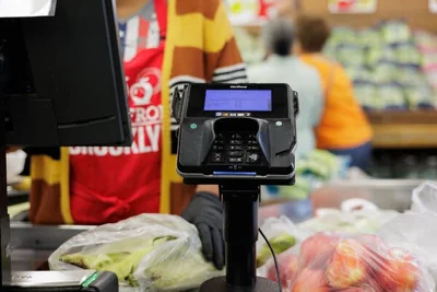 A cashier scans produce with a credit card terminal in the foreground and shoppers in the background.