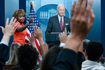 President Biden stands at a podium. A woman in a bright orange dress stands next to him. People seated in front of them raise their hands.