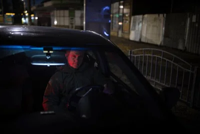 A man sits inside a car at night, his face lit but the rest of the car’s interior dark. 