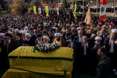 A dense crowed of men, some in white turbans, stand with flags near a coffin draped in a yellow fabric and topped by a large wreath.