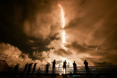 A group of people are silhouetted against  clouds lit by the bright trail of a rocket taking off.