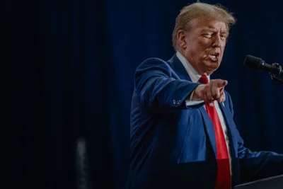 Former President Donald J. Trump speaking at a lectern while wearing a blue suit, white shirt and red tie. He is pointing with one hand. 