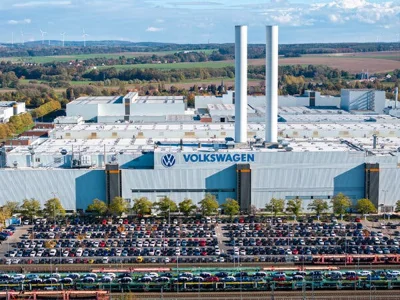 An aerial view of a Volkswagen plant. Lines of cars are parked in a lot outside a building with the company logo.