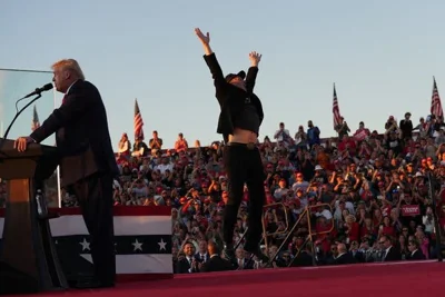 Elon Musk extends his arms upward as he jumps. Donald J. Trump is standing to the stand, speaking into a microphone on a lectern.