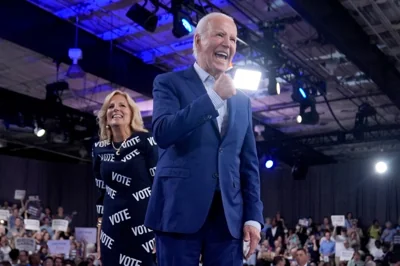Joe Biden and the first lady at a campaign rally in North Carolina. He is in a blue suit and looks animated. She is wearing a black dress with the words VOTE written all over it. 