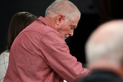 A gray-haired man in a red shirt looks down at a court hearing.