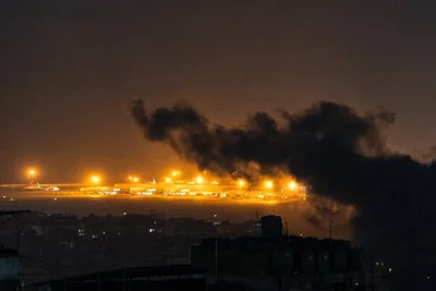 A nighttime photograph showing an airport in the distance, with a cloud of smoke in the foreground.