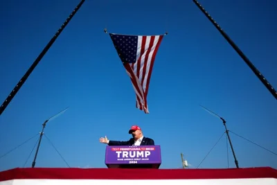 Former President Donald J. Trump, in a dark jacket and a red cap, speaks in Butler, Pa., beneath an American flag. The sign beneath  his microphone says “TRUMP: Make America Great Again.”