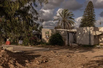 A shot of the exterior of a few small buildings, with pine and palm trees in the background. Mounds of dirt are in the foreground.