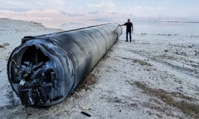 A ballistic missile lies on the shore of the Dead Sea – a section of a huge pipe-shaped metal object is seen on the sand, its diameter and height near that of a man standing next to it; circuitry and mechanical components spill out of the end of it; it is dark-coloured and smoke-damaged, and appears in contrast to the pale sand, sea and pale blue sky