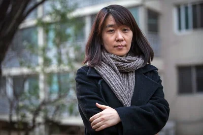 A woman in a black jacket and brown scarf stands with her arms folded, looking straight at the camera.