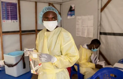 A health worker in a yellow gown, a white mask and a blue hairnet holds a sealed plastic bag containing samples in a makeshift laboratory space in a tent.