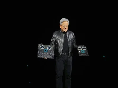 Jensen Huang, in a black leather coat and black clothing, holds two computer chip displays.