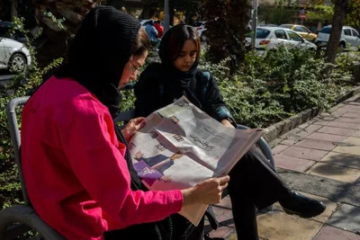 Two women sit on a bench, one reading a newspaper.