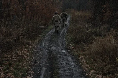 Under overcast skies, two soldiers walk away from the camera on a narrow muddy road with brush on either side.