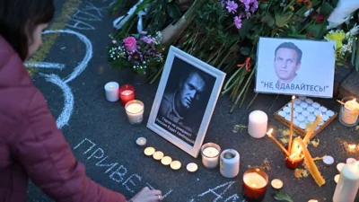 A woman uses chalk to write a message by a portrait of Alexey Navalny displayed amongst flowers and lit candles as protesters pay their respects outside the Russian Embassy in Belgrade, on February 16, 2024, following the news of Russian opposition leader and political activist Alexey Navalny's death at the Arctic prison where he was serving a 19-year-term. (Photo by OLIVER BUNIC / AFP)