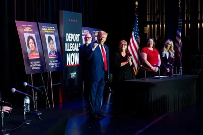 Donald Trump stands onstage holding his fist up. There are signs behind him and American flags. He is wearing a blue suit and red tie.