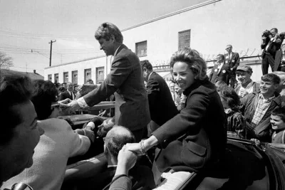 A black and white photo of Mrs. Kennedy sitting on top the back seat of a convertible automobile on a sunny day as she shakes  a well wisher’s hand while her husband stands in the well of the car also shaking hands. 