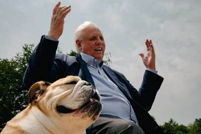 A man with white hair and a blue suit jacket raises his arms. A bulldog sits next to him.