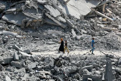 A woman and a young girl walk on a dusty path cutting between enormous mounds of rubble. 