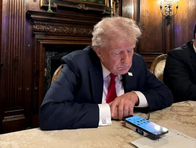 Donald Trump sits at a desk next to another man and looks at his phone, which shows the X Space.